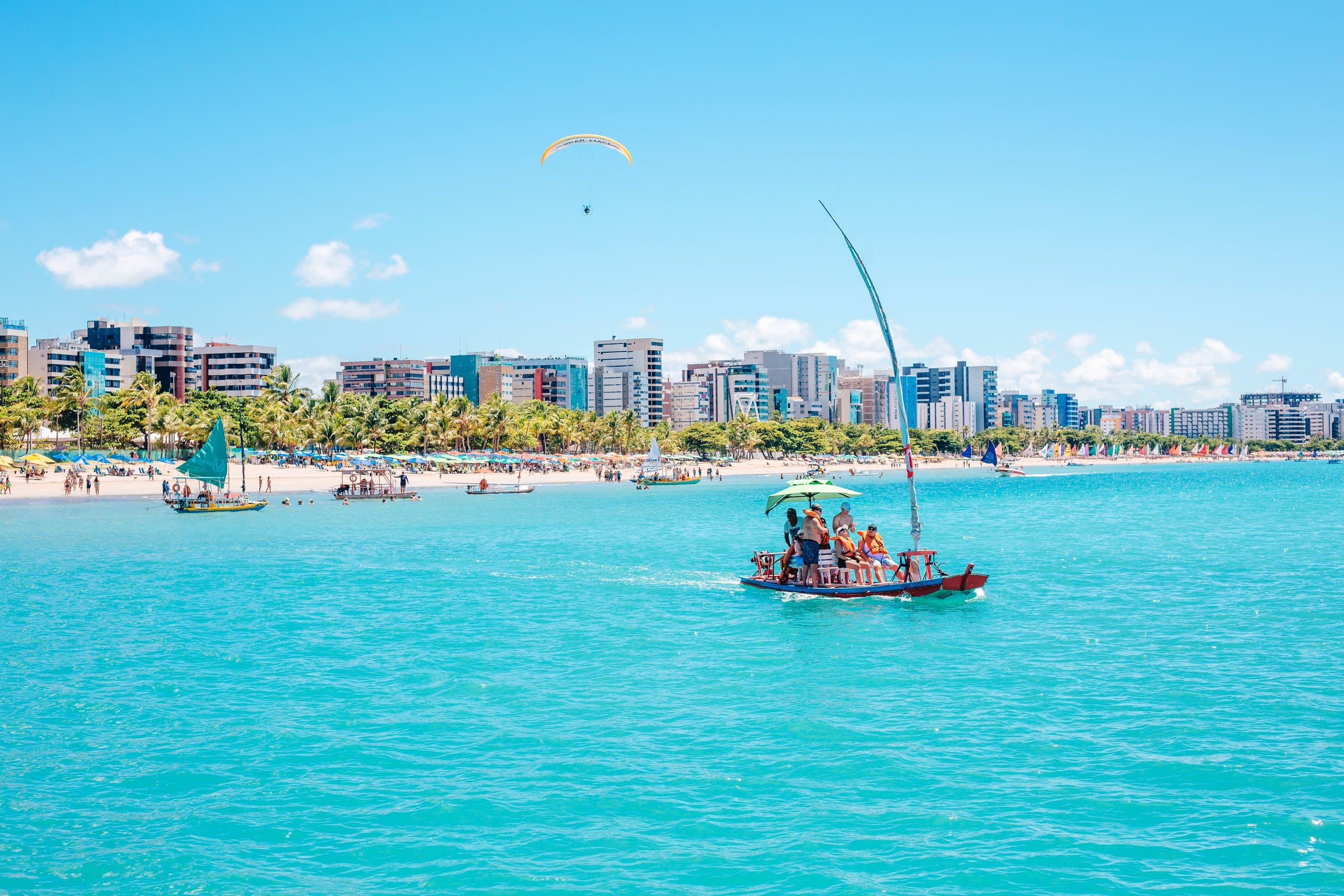 Jangada levando passageiros no mar turquesa de Maceió, com a faixa de prédios da orla ao fundo